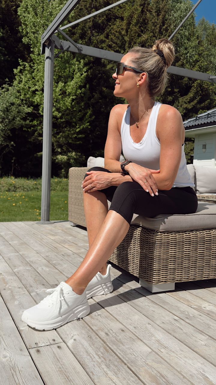 Kvinne i hvit treningssinglet, svarte treningsbukser og hvite joggesko sitter på en flettet hagebenk på en treterrasse utendørs med grønn hage og trær i bakgrunnen.