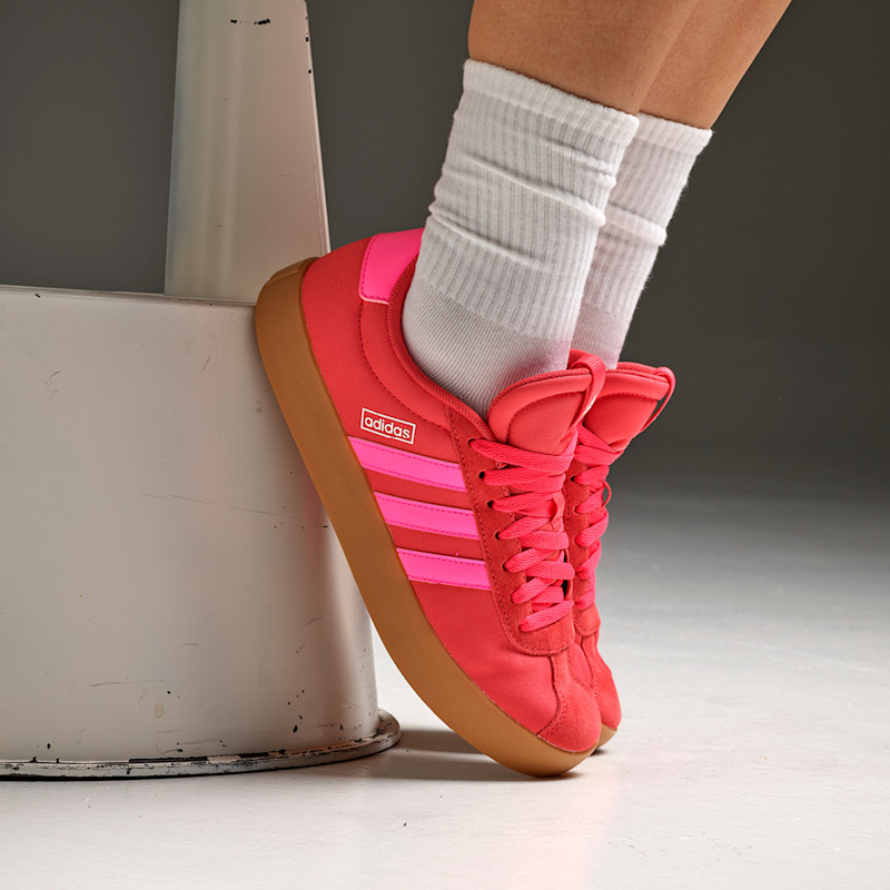 Rød Adidas sneaker med rosa striper og gummisåle, brukt med hvite sokker mot en lys bakgrunn.