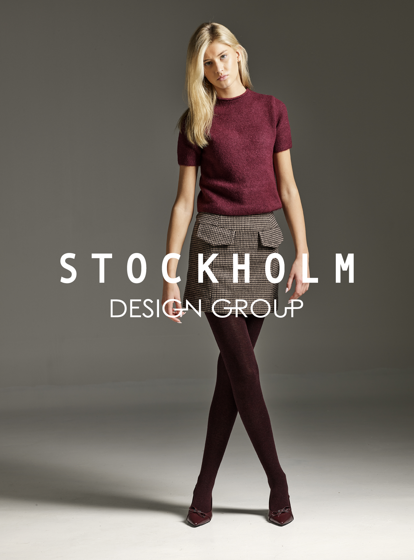 Motemodell i burgunder genser og rutet skjørt med matchende strømpebukser og spisse, vinrøde pumps med dekorativ spenne.