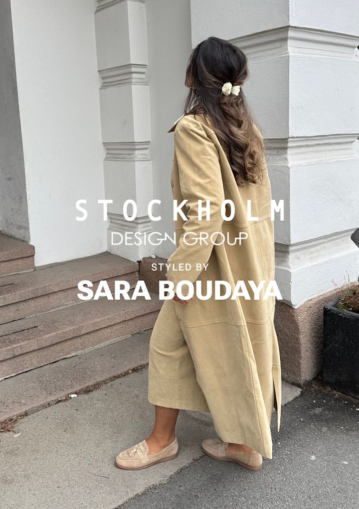 Kvinne med langt, bølget hår pyntet med hvite blomster går opp trapp utenfor bygning iført beige lang kåpe og sko.