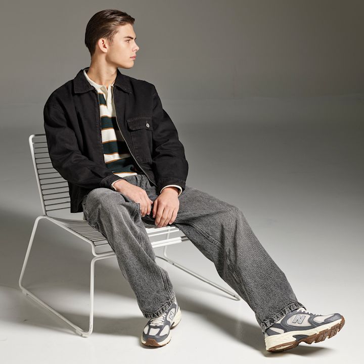 Gutt sittende på hvit stol med svart jakke, stripete genser og grå jeans, iført grå og hvite New Balance sneakers i retrostil.