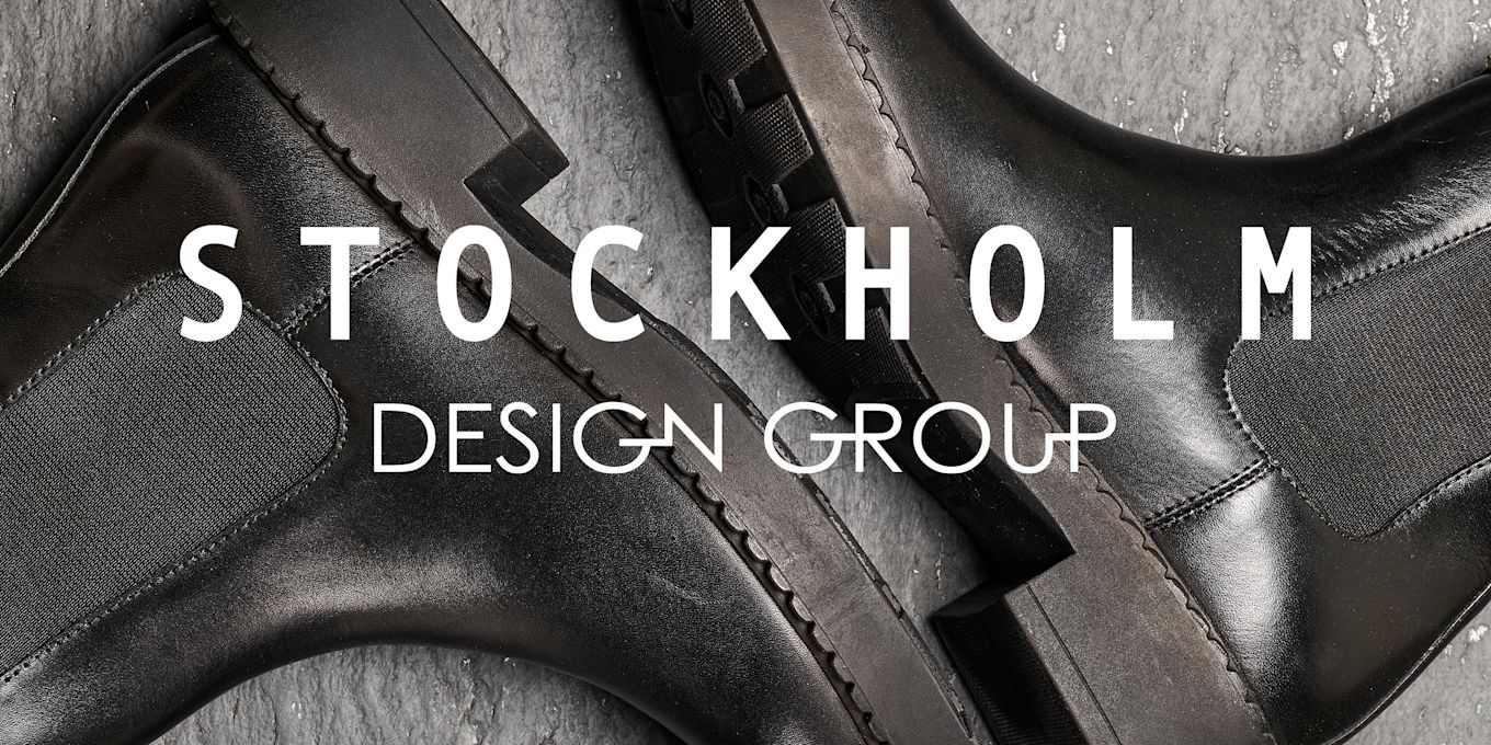 Elegante svarte skinnsko fra Stockholm Design Group på grå bakgrunn, med stilrene detaljer og solid såle. Hvit logo over.