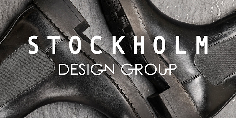 Elegante svarte skinnsko fra Stockholm Design Group på grå bakgrunn, med stilrene detaljer og solid såle. Hvit logo over.