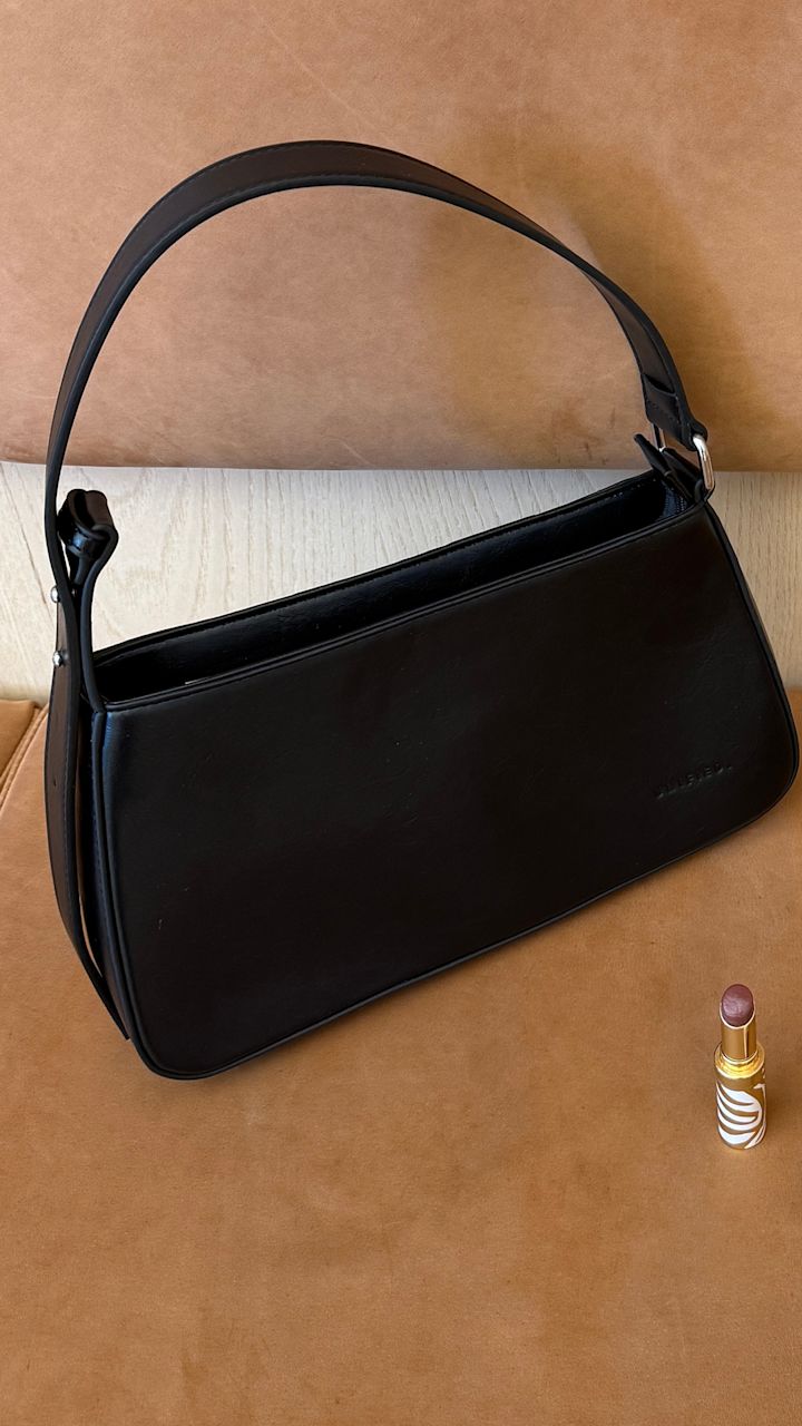 Elegant svart skinnveske fra Unified med håndtak på brun bakgrunn, med en leppestift i gull og hvitt ved siden av.