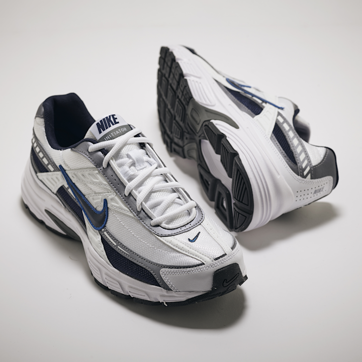 Hvite og grå Nike Initiator joggesko med blå detaljer, plassert på hvit bakgrunn, med den ene skoen liggende på siden som viser sålen.