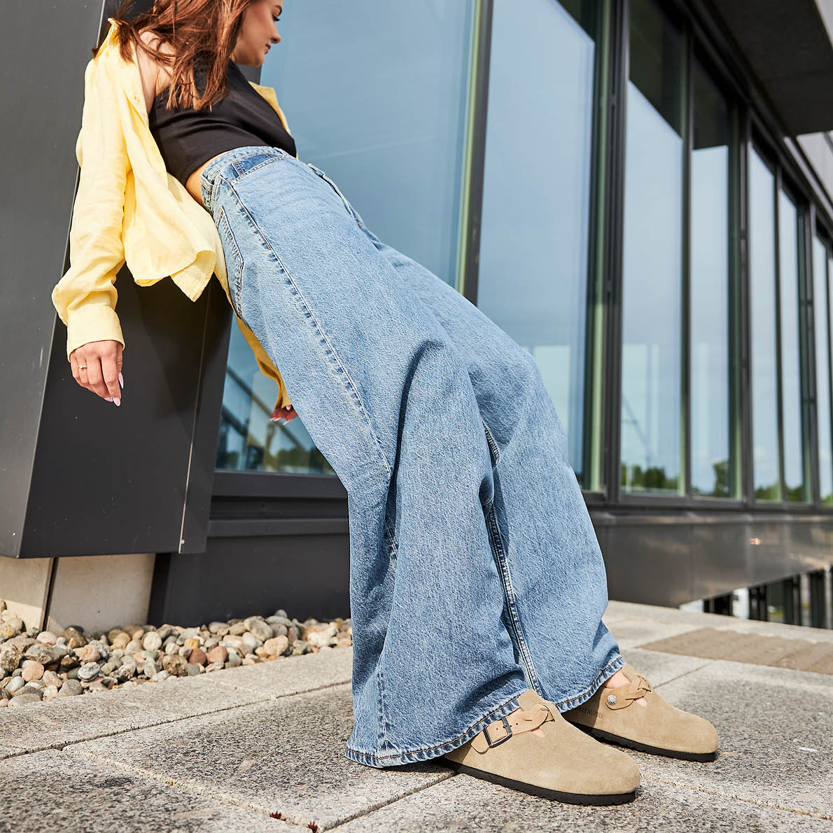 Kvinne i vid jeans og gul skjorte lener seg mot vegg, med beige semskede sandaler med spenne i casual stil.