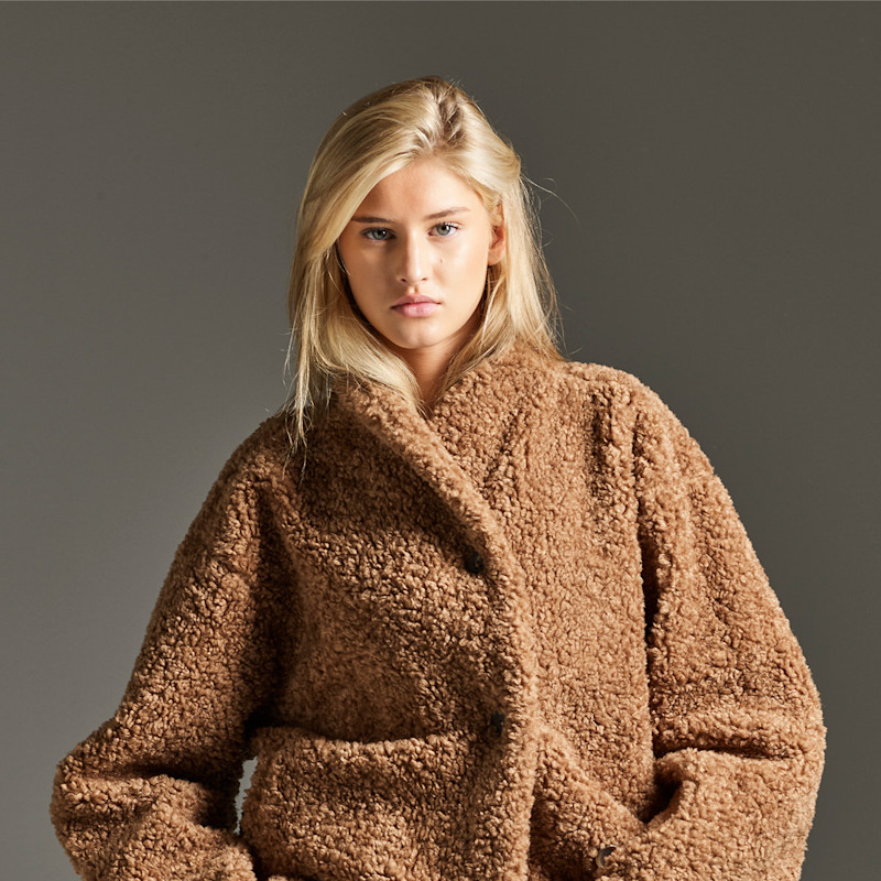 Jente med langt blondt hår iført en brun, fluffy teddyjakke mot en grå bakgrunn.
