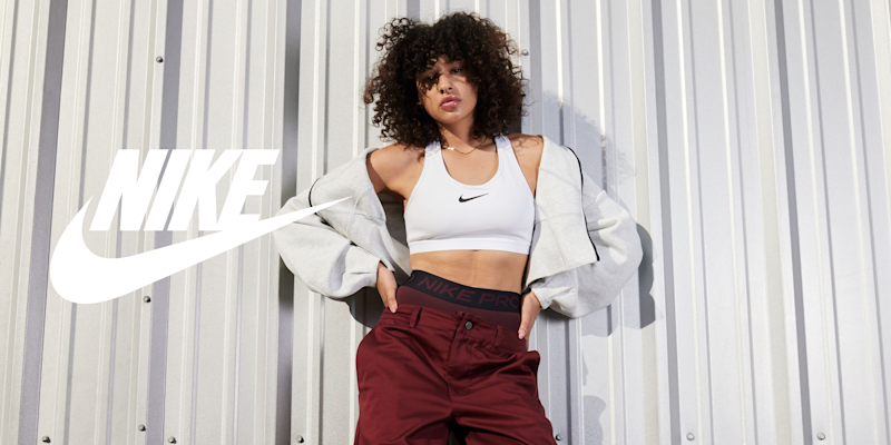 Kvinne i hvit Nike sports-BH, grå jakke og burgunder bukser poserer foran en metallvegg med Nike-logo til venstre.