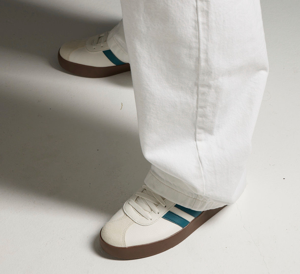 Hvite sneakers med turkise striper og brun såle, brukt med hvite bukser på en lys bakgrunn.