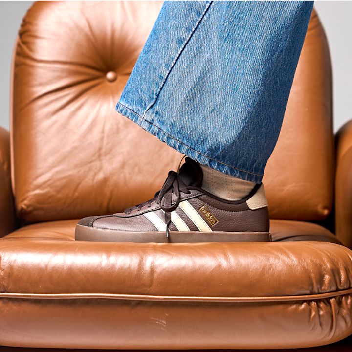 Mørkebrun Adidas-sneaker med kremhvite striper og gulldetaljer, stylet med blå jeans på en brun lærsofa.
