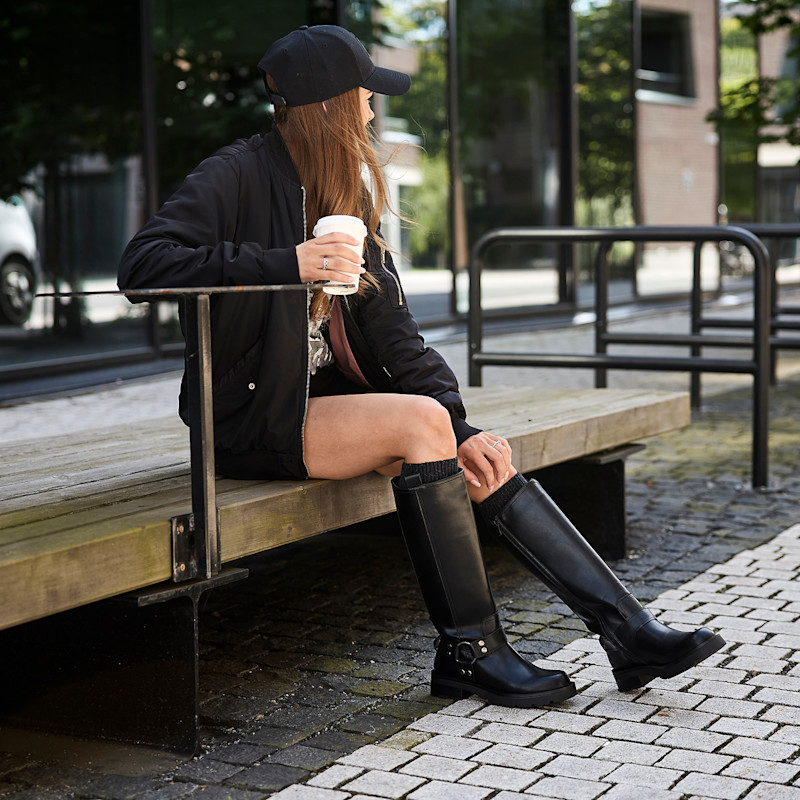 Ung kvinne i svart caps, jakke og høye svarte støvler sitter på benk utendørs og holder en kaffekopp.