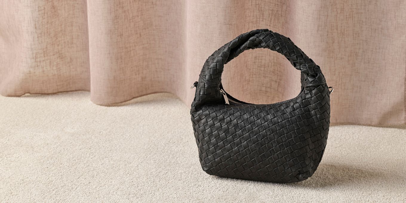 Svart flettet skinnveske med buet håndtak på beige teppe foran lyse gardiner.
