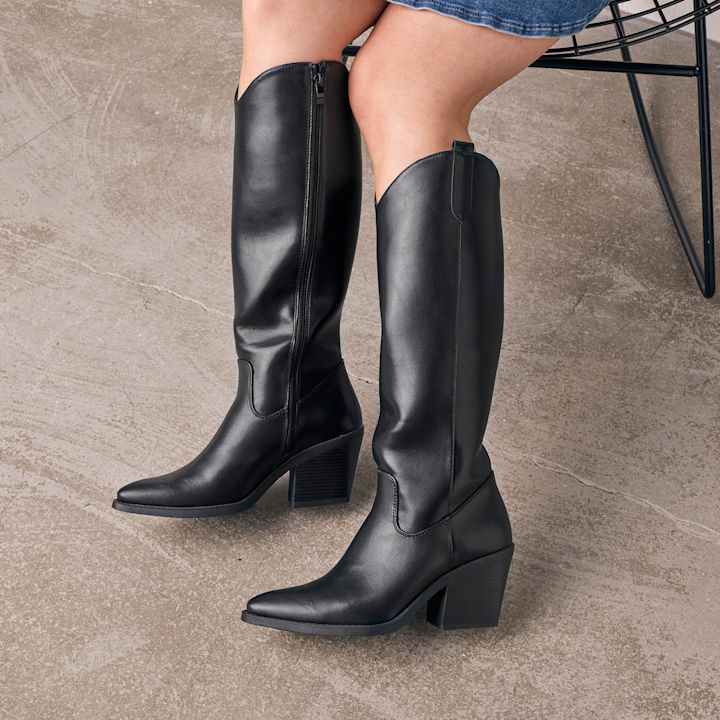 Sorte skinnstøvler med høye hæler og spiss tupp, båret av en person som sitter på en stol med betonggulv i bakgrunnen.