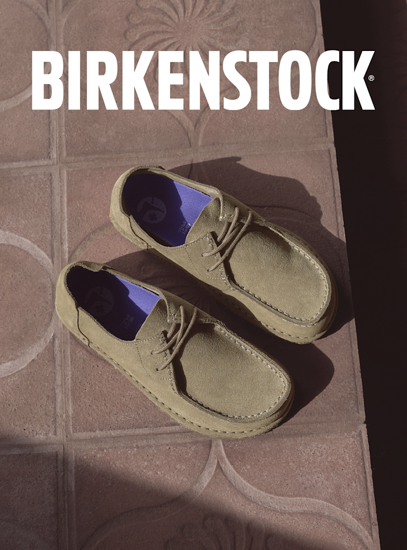 Beige semsket Birkenstock Utti med blått innerfôr på flislagt gulv, med Birkenstock-logo øverst i bildet.