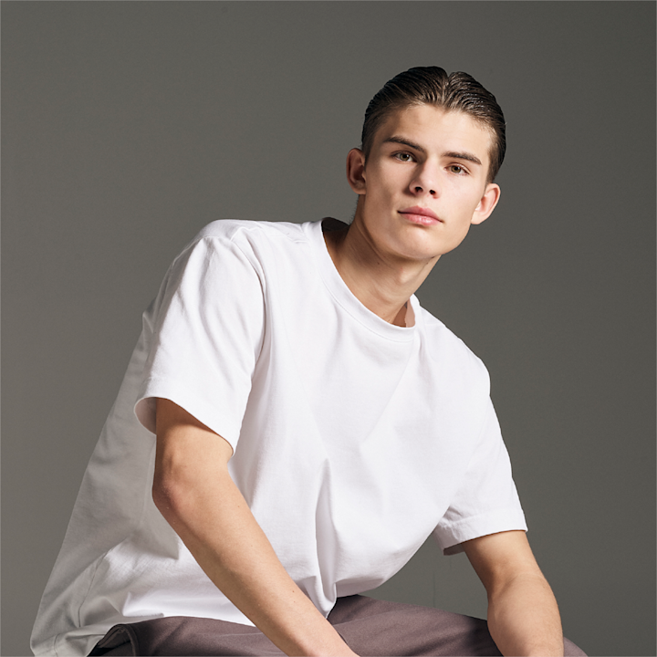 Ung mann med mørkt, gredd hår som sitter og ser rett fram, iført en hvit t-skjorte og mørke bukser mot en grå bakgrunn.