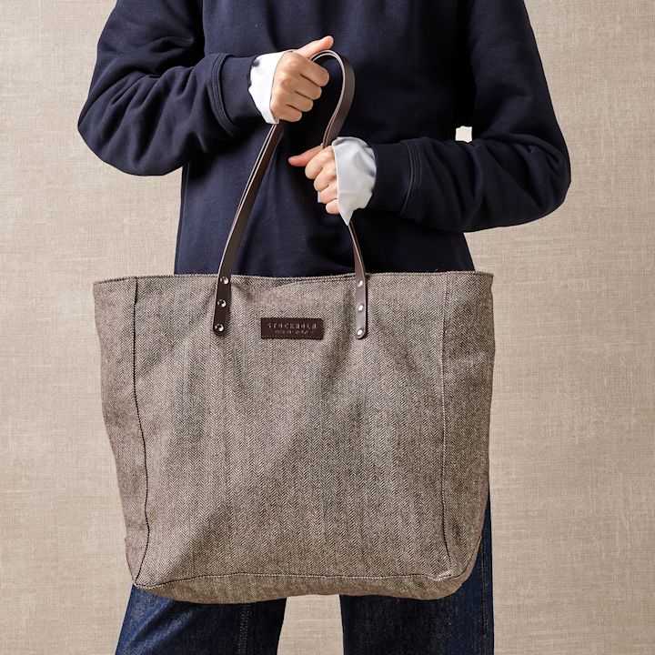 Person i mørkeblå genser holder en stilfull gråbrun stoffveske med lærhåndtak mot en nøytral beige bakgrunn.