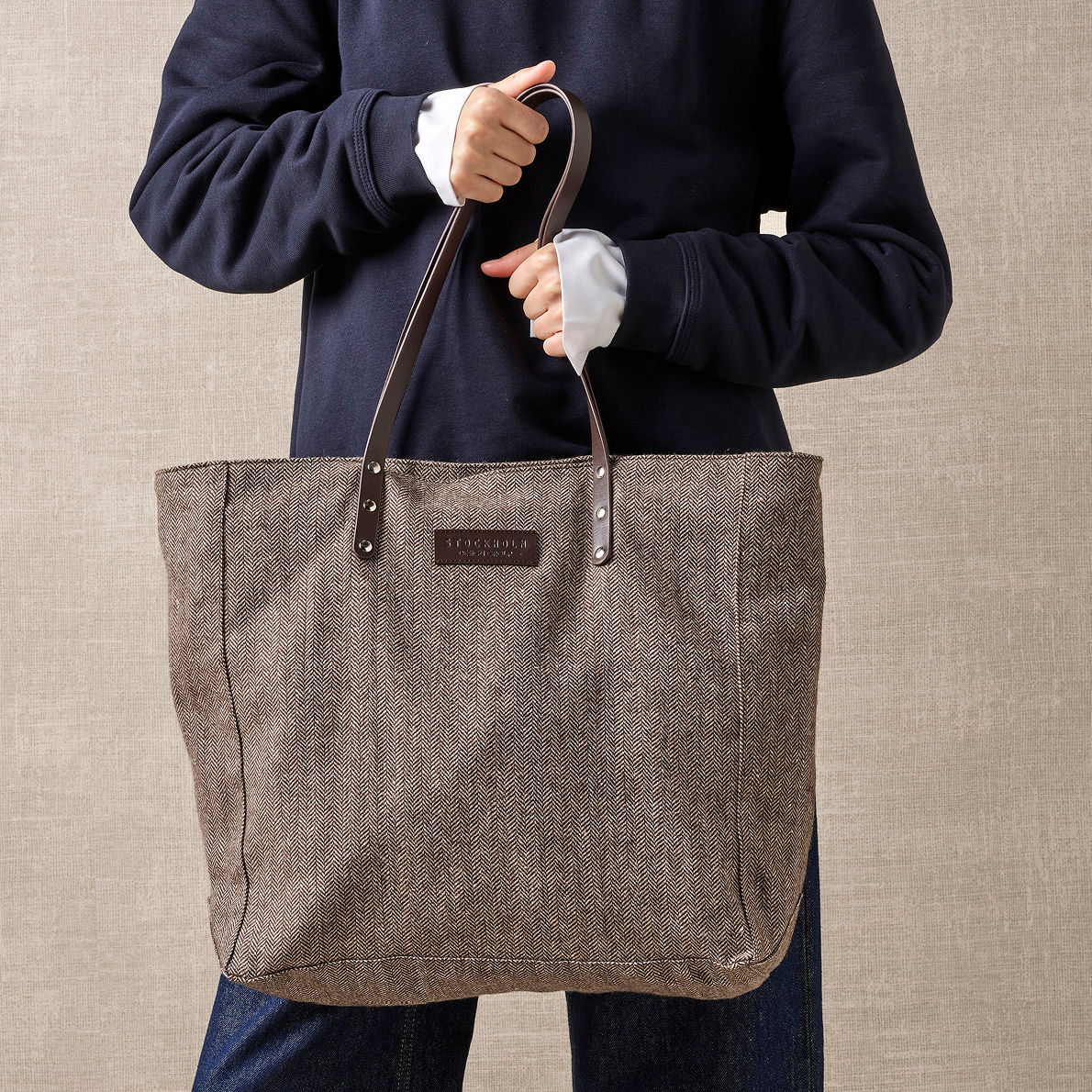 Person i mørkeblå genser holder en stilfull gråbrun stoffveske med lærhåndtak mot en nøytral beige bakgrunn.