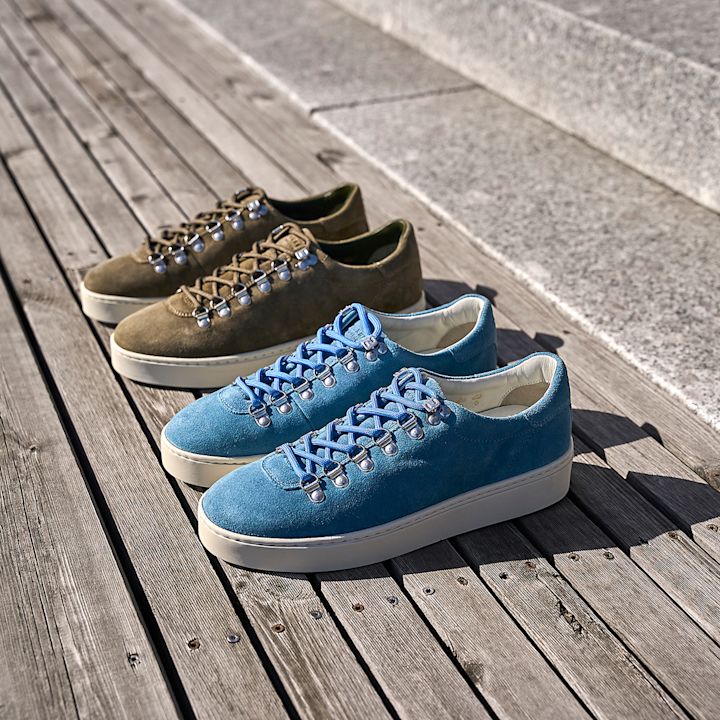 To par sneakers i semsket skinn: én blå og én olivengrønn, begge med metallhemper og hvit såle, på et tredekke.