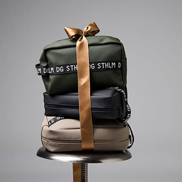 Tre toalettmapper i mørkegrønn, svart og beige, stablet oppå hverandre og bundet sammen med et gullfarget bånd.