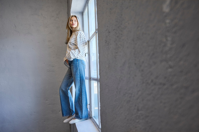 Jente i stripete genser og jeans står ved vindu, iført hvite, enkle sneakerinas i minimalistisk stil.