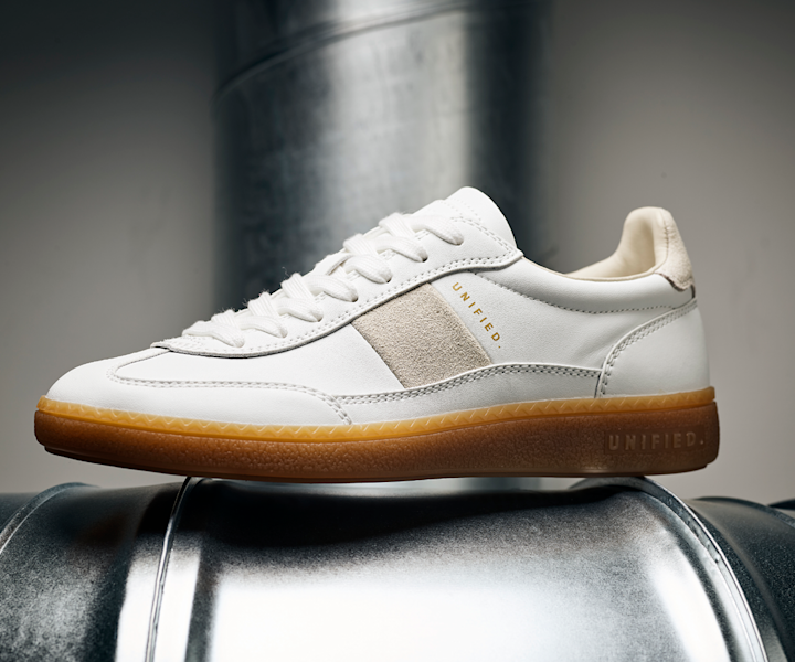 Hvit sneaker med beige semsket detalj og brunt gummisåle, plassert på en metalltønne i et nøytralt miljø.