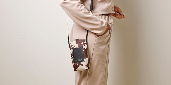 Person i beige antrekk med liten crossbody-veske i ku-mønster, hvit og brun med sort lomme og stropp.