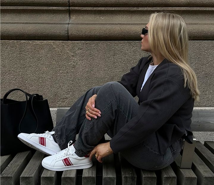 Kvinne med blondt hår og solbriller sitter på en benk ved en steinvegg, iført svart jakke, grå bukser og hvite sneakers med røde detaljer, med en svart veske ved siden av seg.