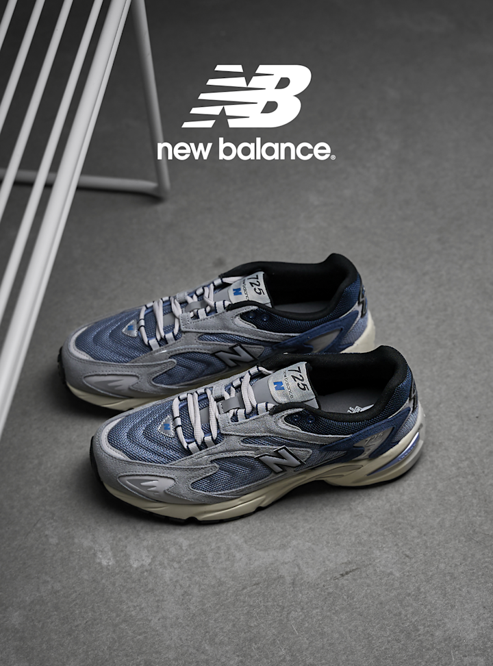 New Balance 725 løpesko i blå og grå farger med hvite lisser, plassert på grått underlag med New Balance-logo øverst.