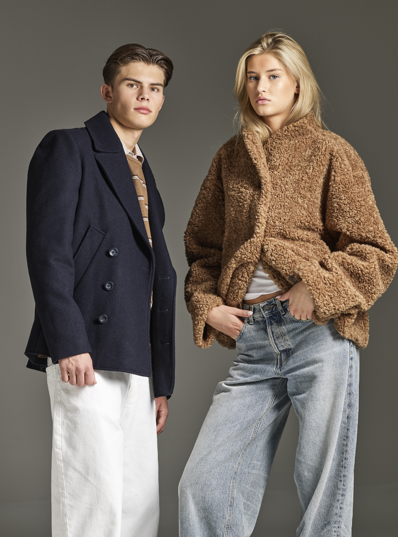 To personer poserer i moteantrekk - en i mørkeblå jakke med hvite bukser og en i brun teddy-jakke med lyse jeans.