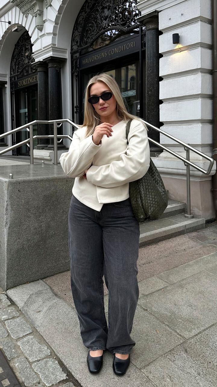 Kvinne med solbriller, hvit genser, grå bukser og svart veske står foran inngang med skiltet "Halvorsens Conditori" i bymiljø.