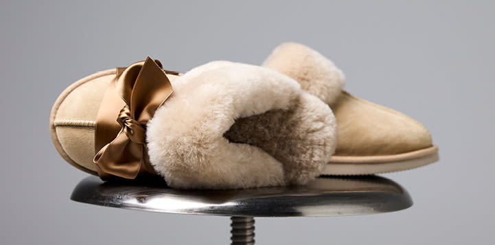 Beige tøfler med myk pelskant, plassert på en metallisk overflate mot grå bakgrunn, med et gyldent silkebånd rund den ene tuppen.
