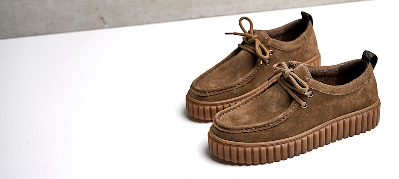 Brune semsket mokasiner med snøring og tykk gummisåle i creeper-stil, plassert på hvit overflate.