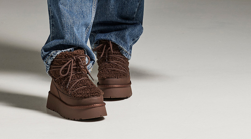 Brune, lodne ankelstøvletter med plattformsåle og snøring, brukt sammen med blå jeans på grå bakgrunn.