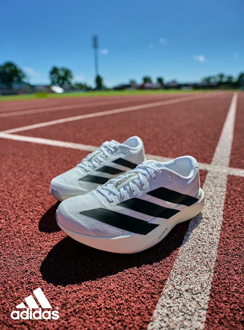 Hvite Adidas Adizero løpesko med svarte striper på rød friidrettsbane under blå himmel.