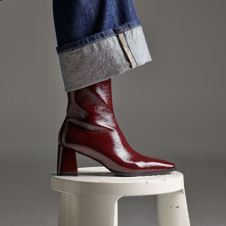 Vinrød lakkankelstøvlett med blokkert hæl, spiss tå, båret med oppbrettede jeans på hvit krakk.