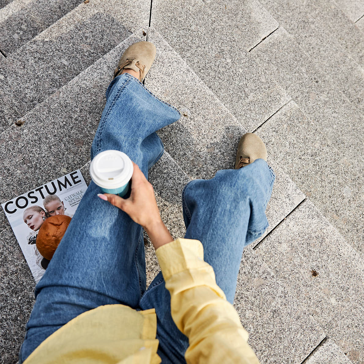 Kvinne med beige semskede sandaler og blå jeans sitter på steintrapp, holder kaffekopp og moteblad i hånden.