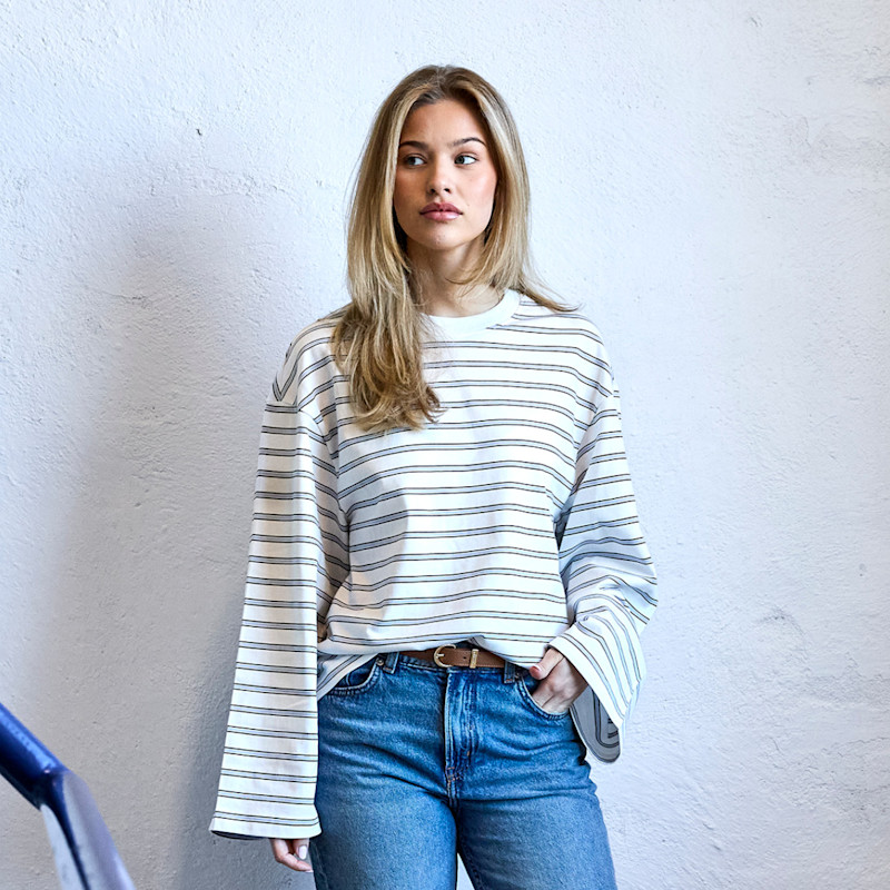 Ung kvinne med blondt hår iført hvit stripete genser med vide ermer og blå jeans med brunt belte.