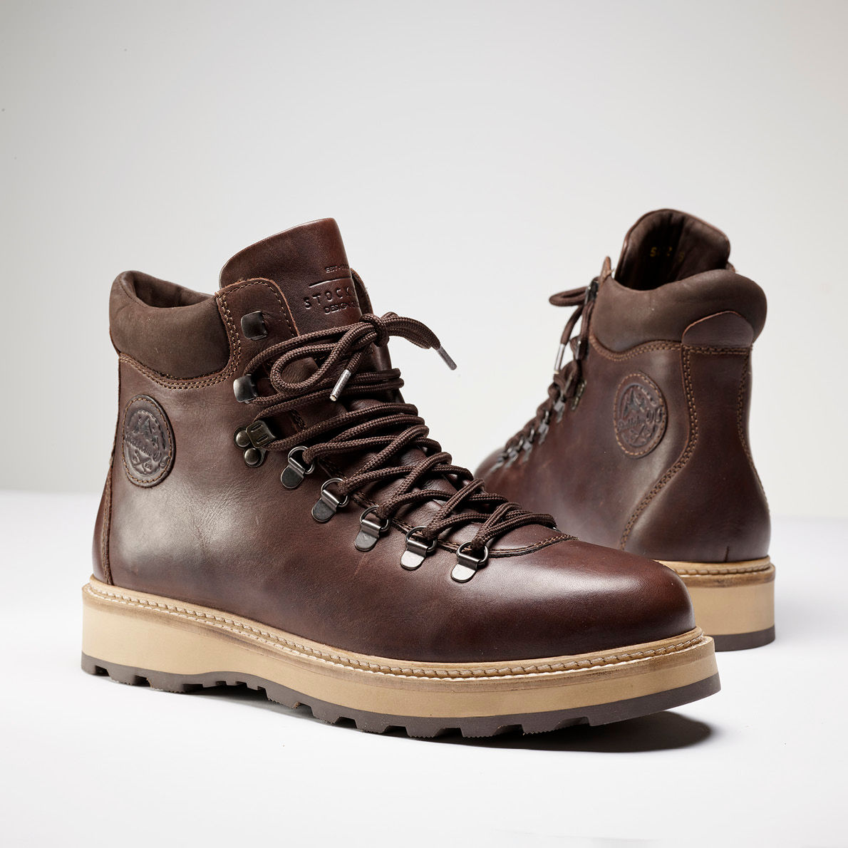 Brune lærstøvler i fjellstil med snøring, robuste såler i beige og merkelogo på siden, mot hvit bakgrunn.