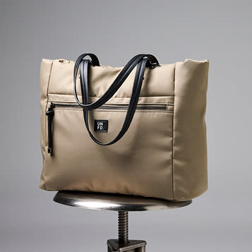 Beige stoffveske med svarte håndtak og glidelåslomme, plassert på en metallkrakk mot grå bakgrunn.