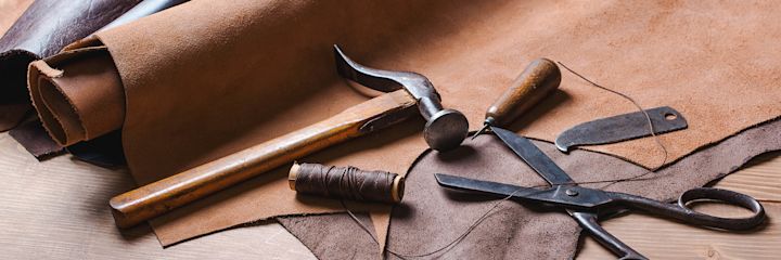 Rull med lær, hammer, trådsneller, skreddersaks og lærverktøy plassert på et bord med lærstykker.