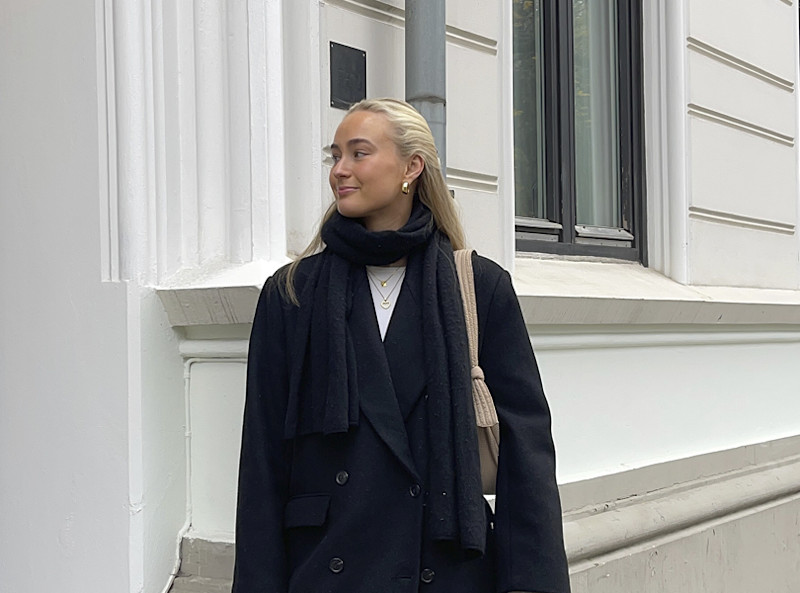 Ung kvinne iført svart kåpe og skjerf står og ser til siden foran hvit bygning med vindu.