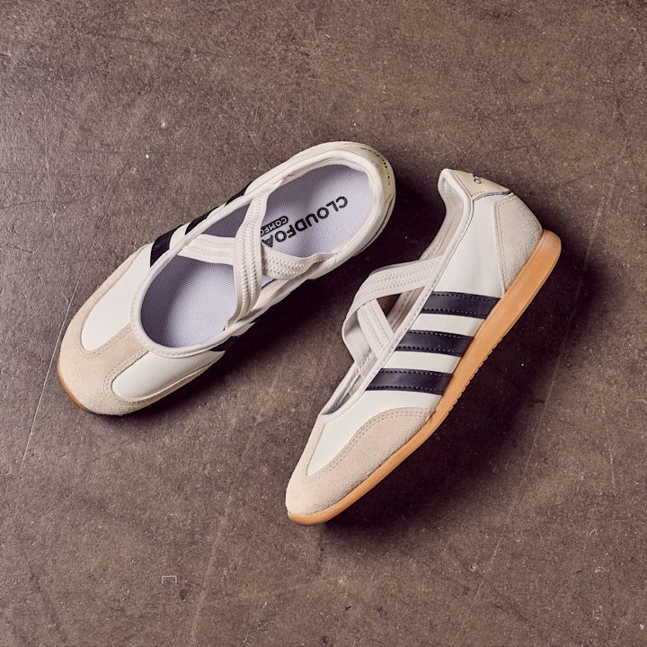 Hvite adidas sneakerinas med svarte striper, elastiske remmer og gummibunn, plassert på mørk betongoverflate.