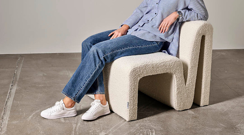 Person sitter på en moderne, kremfarget stol med bølget design i et rom med betonggulv.