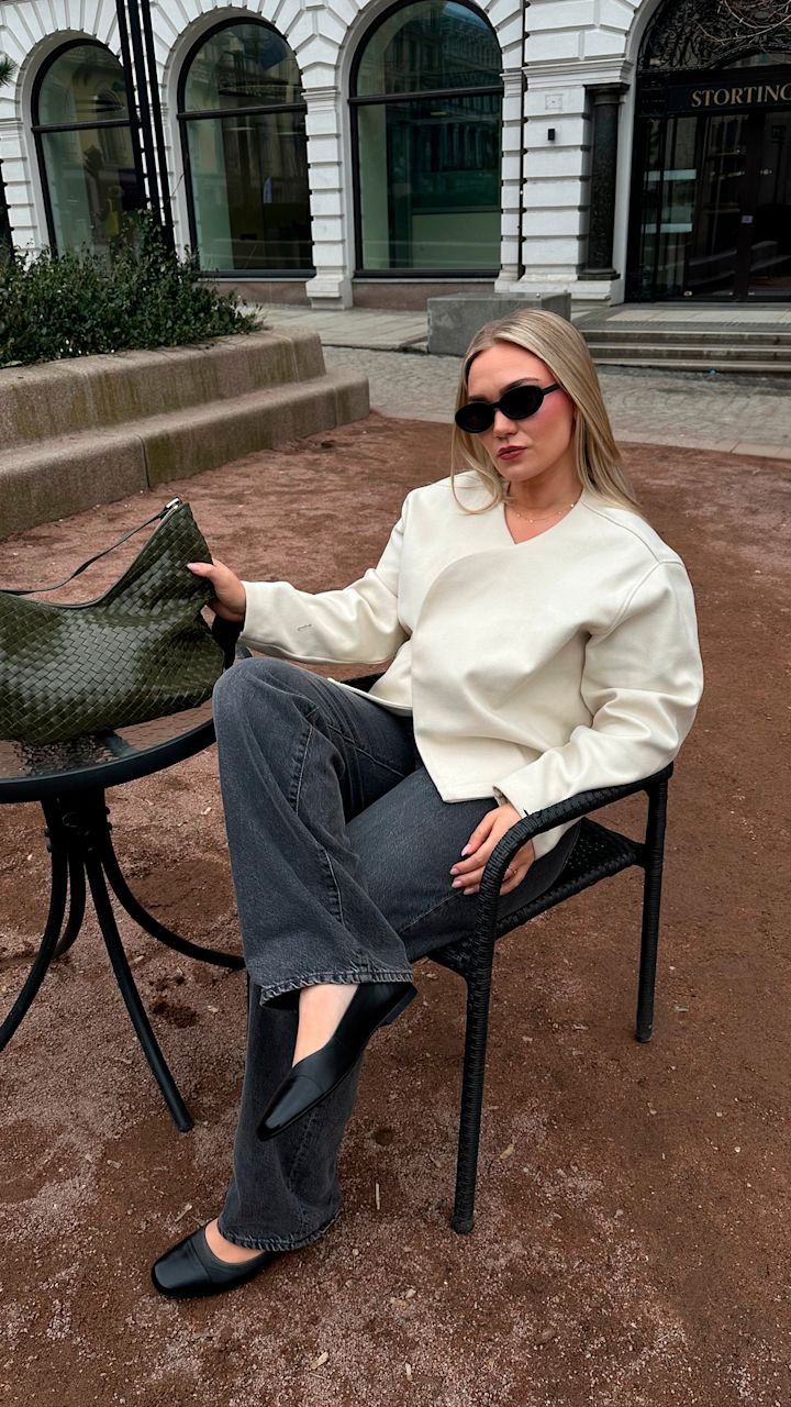 Kvinne med solbriller sitter i svart kurvstol utendørs, iført hvit jakke, mørke jeans og svarte sko, med grønn veske på glassbord ved siden av.