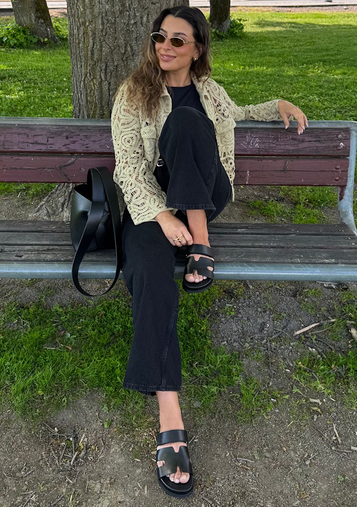 Influenser Sara Boudaya på parkbenk med kremfarget heklet jakke og svart antrekk, iført svarte og trendy skinnsandaler med remmer.