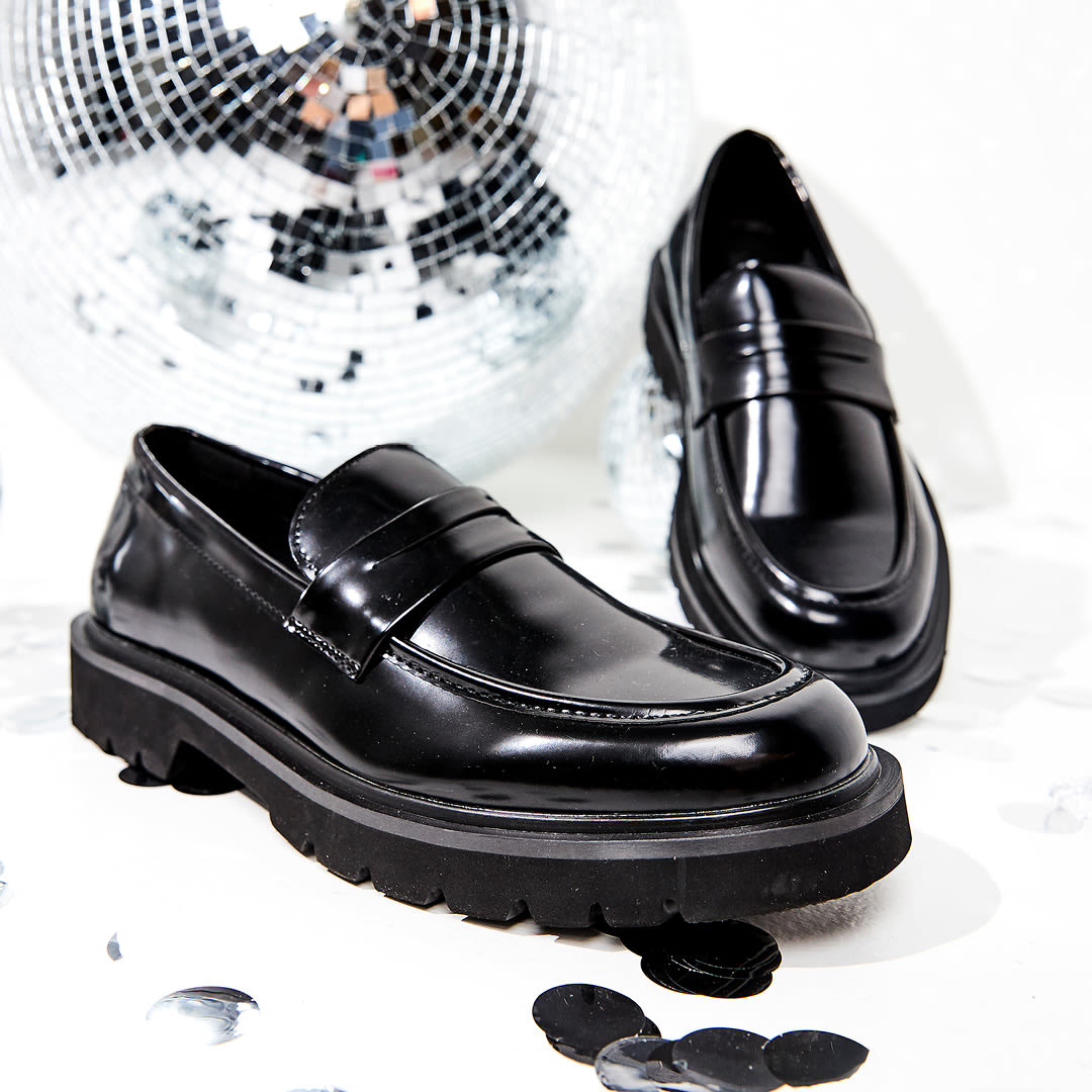 Elegante svarte, blanke loafers med kraftig såle ved siden av en diskokule.