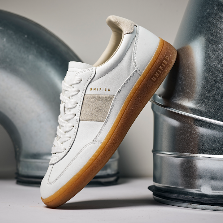 Hvit UNIFIED sneaker med beige detaljer og gummiaktig brun såle, plassert ved metalliske gjenstander.