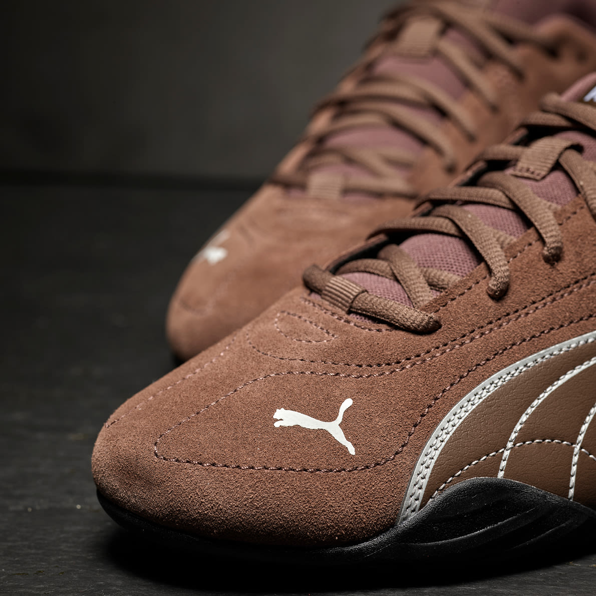 Brune Puma sportssko i semsket skinn med hvite detaljer og svart såle, vist i nærbilde mot mørk bakgrunn.