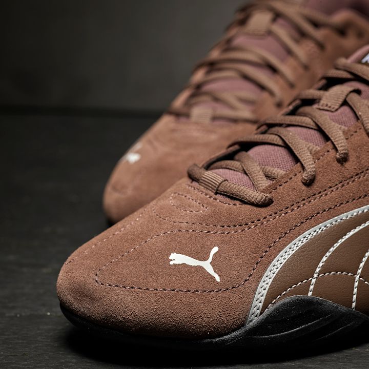 Brune Puma sportssko i semsket skinn med hvite detaljer og svart såle, vist i nærbilde mot mørk bakgrunn.