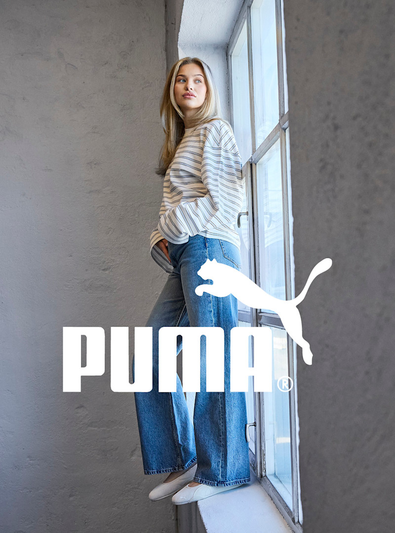 Kvinne i stripete topp og vide jeans med hvite Puma ballerinasko, lener seg mot et vindu. Puma-logo synlig.